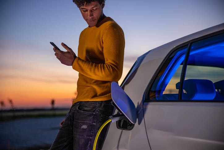 African American man using mobile phone while he is waiting for his electric vehicle to be charged at charging station