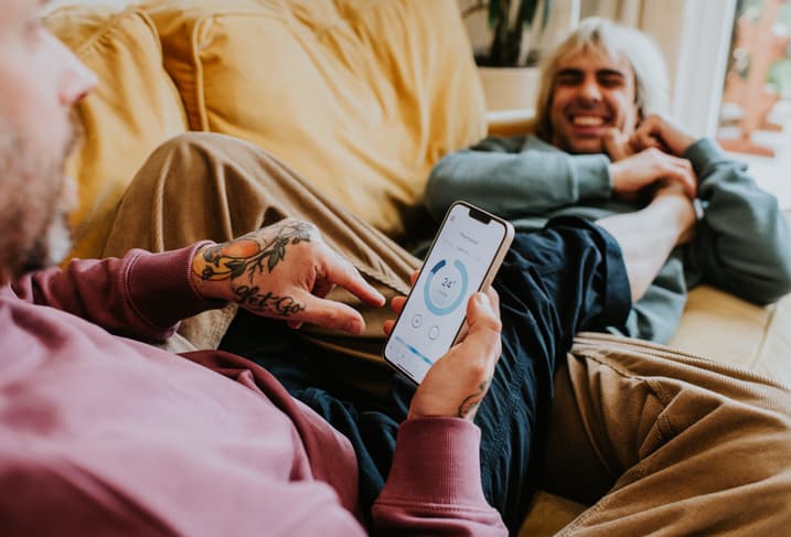 A relaxed couple recline on a soft yellow sofa. One man uses a mobile app to control the home thermostat.